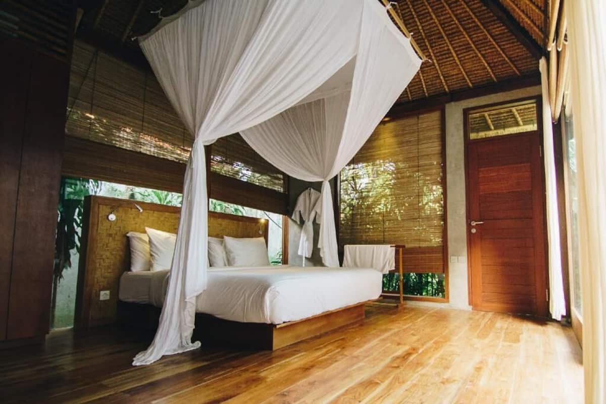 A room with a bed, a wooden floor, a wooden door, and a window with a view of greenery.