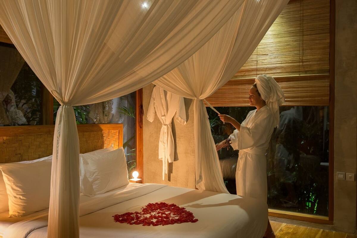 A bedroom with a canopy bed, a bedside table with a lamp, and a view of the outdoors.