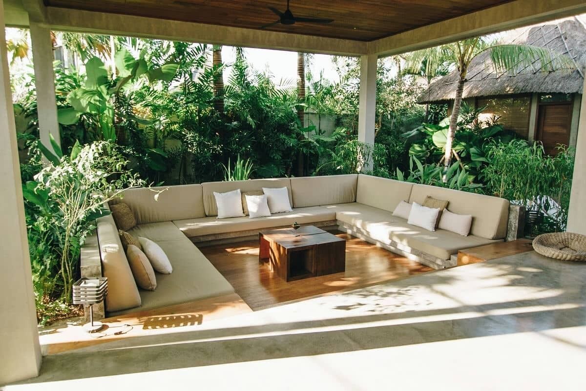 A covered outdoor seating area with a beige sectional sofa, a wooden coffee table, and a thatched roof structure.
