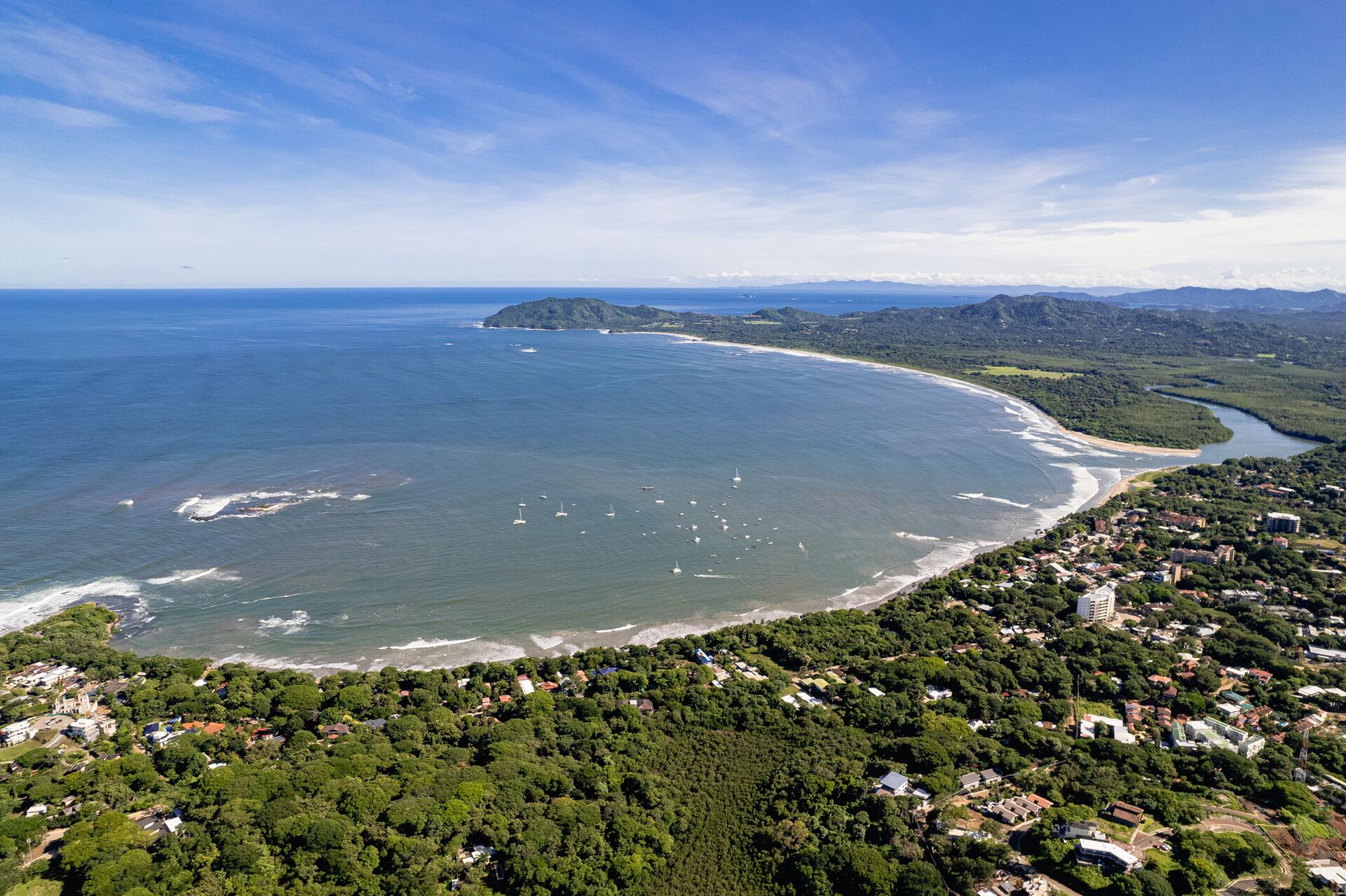 Vue aérienne d’une ville côtière avec une plage, une végétation luxuriante et des bâtiments dispersés.