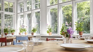 A dining area with a wooden table set for four, surrounded by large windows with potted plants. - Hotel Hornbækhus (Hornbaek)