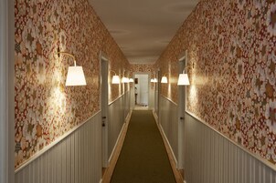 Un pasillo con papel tapiz floral, alfombra verde y lámparas fijadas a la pared.