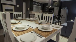 Un coin repas avec une table dressée pour quatre personnes, des chaises blanches et une cuisine en arrière-plan.