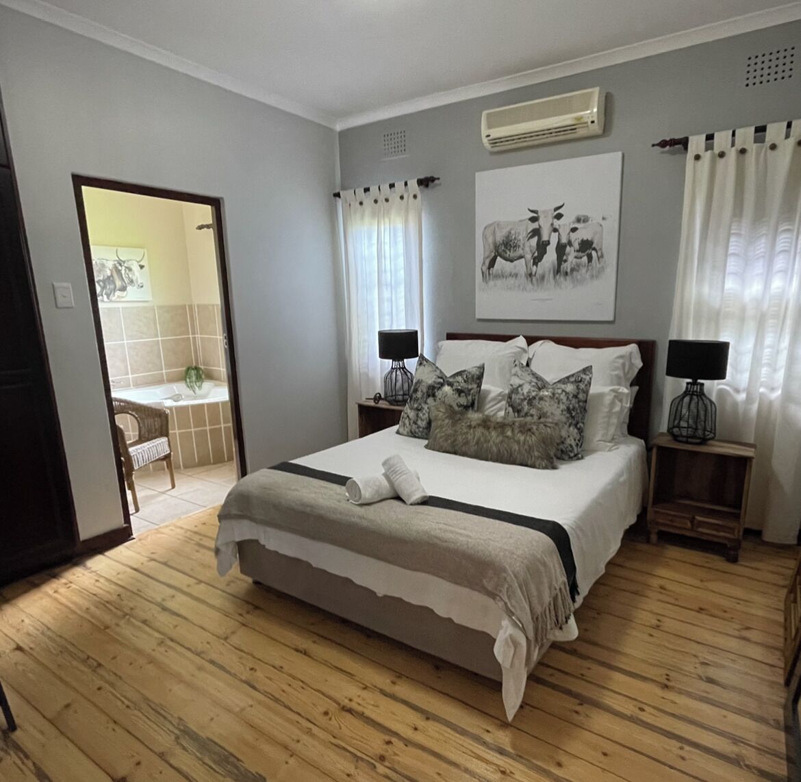 Une chambre avec un grand lit, deux tables de chevet avec des lampes, un climatiseur fixé au mur et une salle de bain visible à travers une porte ouverte.