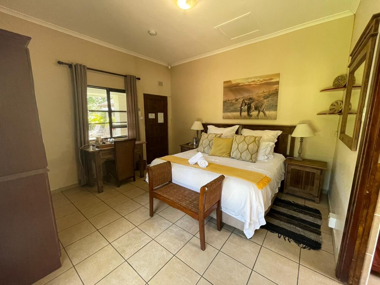 Une chambre d’hôtel avec un lit, un bureau, une chaise, une fenêtre avec des rideaux, un tableau au mur et un petit tapis.