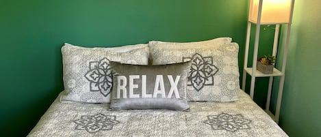 A bed with a patterned bedspread, a pillow with the word "RELAX," and a painting of a beach scene on the wall.