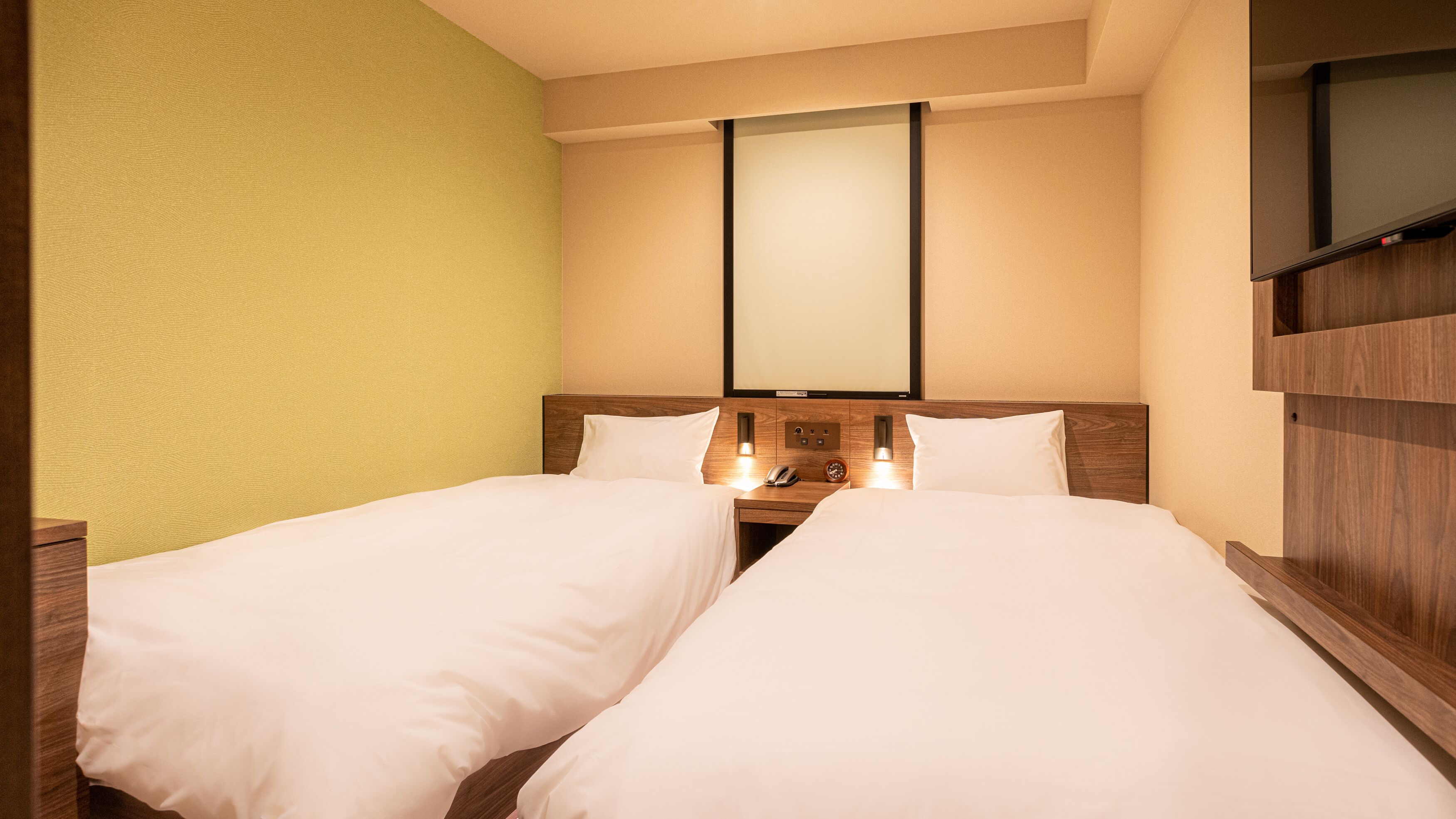 A hotel room with two single beds, a wooden headboard, a bedside table with a phone and a clock, and a wall-mounted TV.