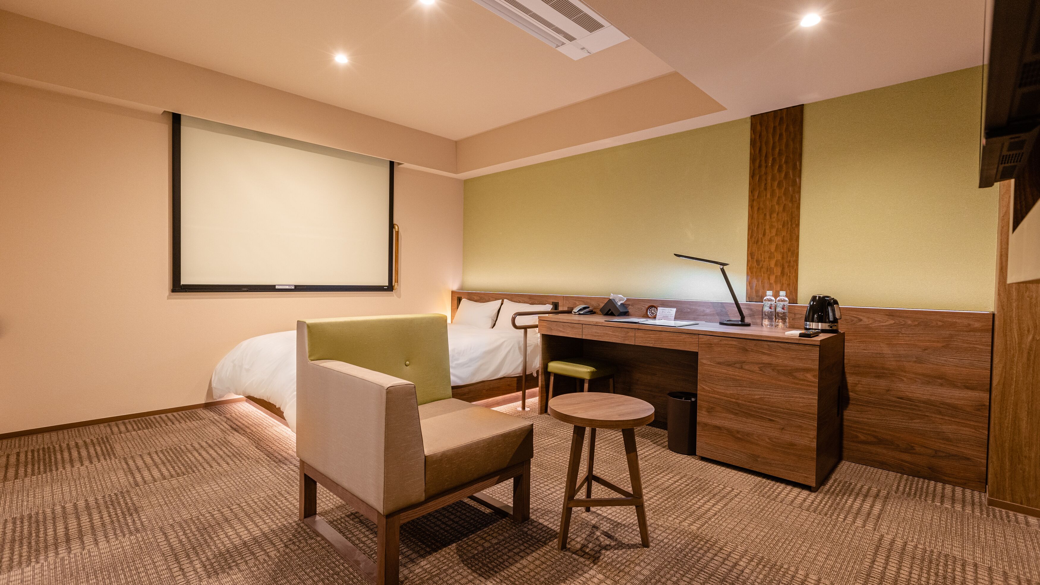 Une chambre d’hôtel comprenant un lit, un bureau, une chaise, un petit tabouret, un écran de projection et un panneau en bois sur le mur.