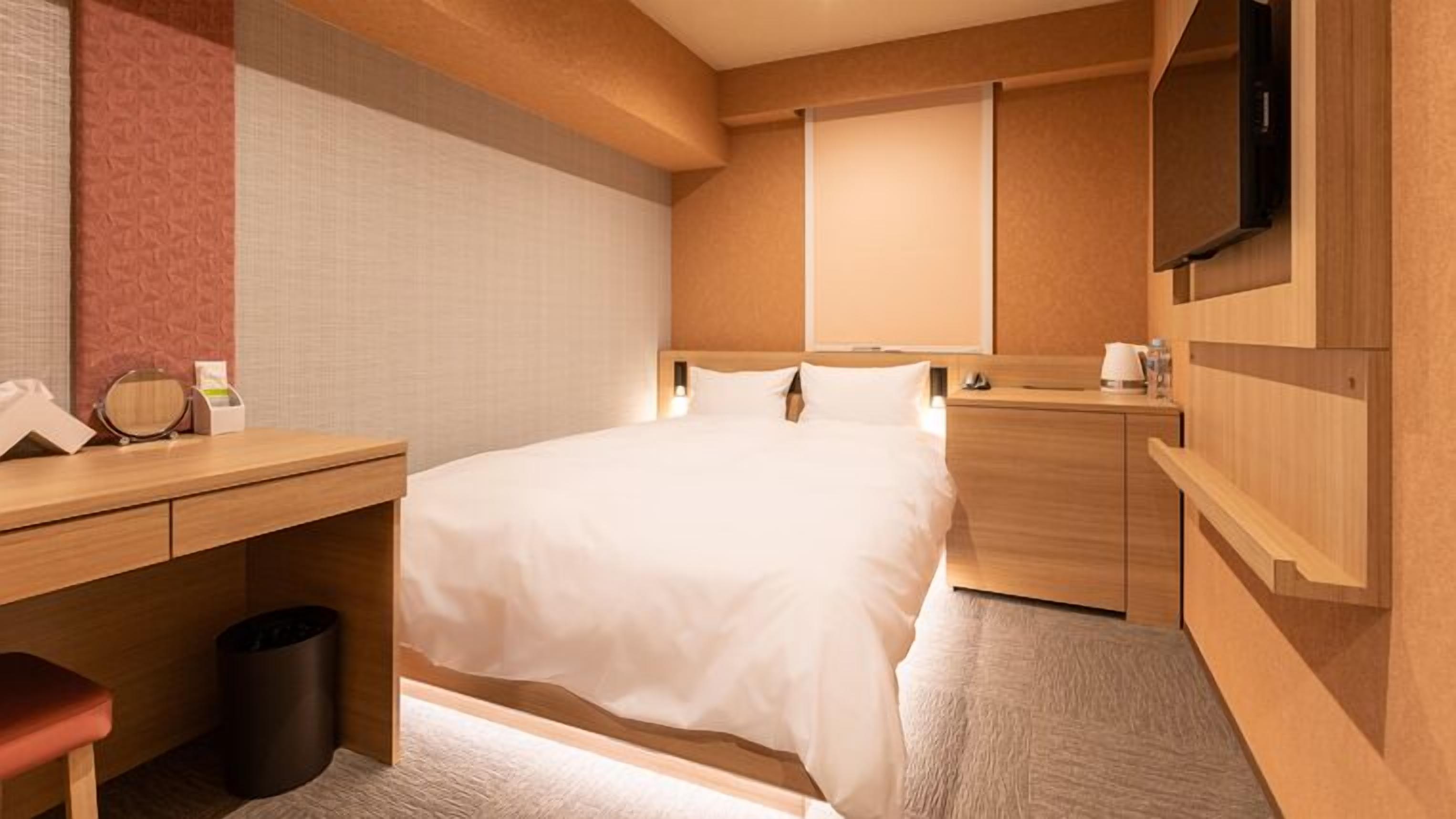 A hotel room with a bed, bedside table, desk, trash bin, and a TV mounted on the wall.