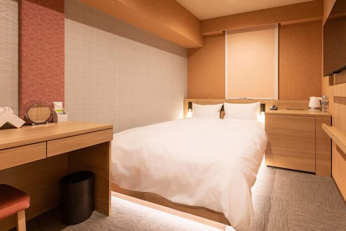 A hotel room with a bed, bedside table, desk, trash bin, and a TV mounted on the wall.