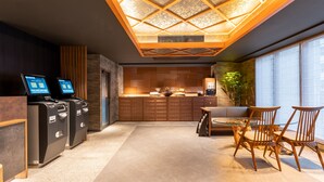 Eine Hotelhalle mit einer Rezeption, Selbstbedienungs-Terminals, einer Sitzecke mit Glastisch und einem großen Fenster mit Jalousien.