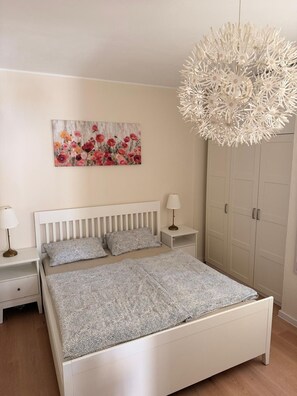Ein Schlafzimmer mit einem weißen Bett, zwei Nachttischen mit Lampen, einem großen Blumenbild an der Wand und einem Kleiderschrank.