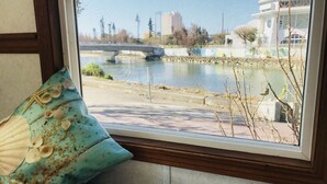 A window with a view of a river, buildings, and a starfish-patterned cushion on a chair. - Mini Chula Inn (Chatan)