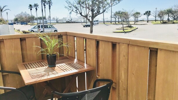 A wooden deck with a table and chairs, overlooking a parking lot with cars and palm trees. - Mini Chula Inn (Chatan)