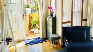 A room with a blue carpet, a black sofa, a wooden table, a small stool, a fan, a potted plant, and a clock on the wall. - Mini Chula Inn (Chatan)