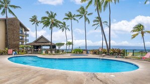 A pool area with palm trees, a building with a stone facade, and a view of the ocean. - SUMMER SALE! Updated 1BR Unit with Pool and Hot Tub Sleeps 4 - Papakea  B201 (Lahaina)
