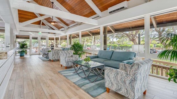 A spacious indoor area with wooden flooring, a high ceiling with exposed beams, and large windows offering views of the outdoors. - SUMMER SALE! Updated 1BR Unit with Pool and Hot Tub Sleeps 4 - Papakea  B201 (Lahaina)
