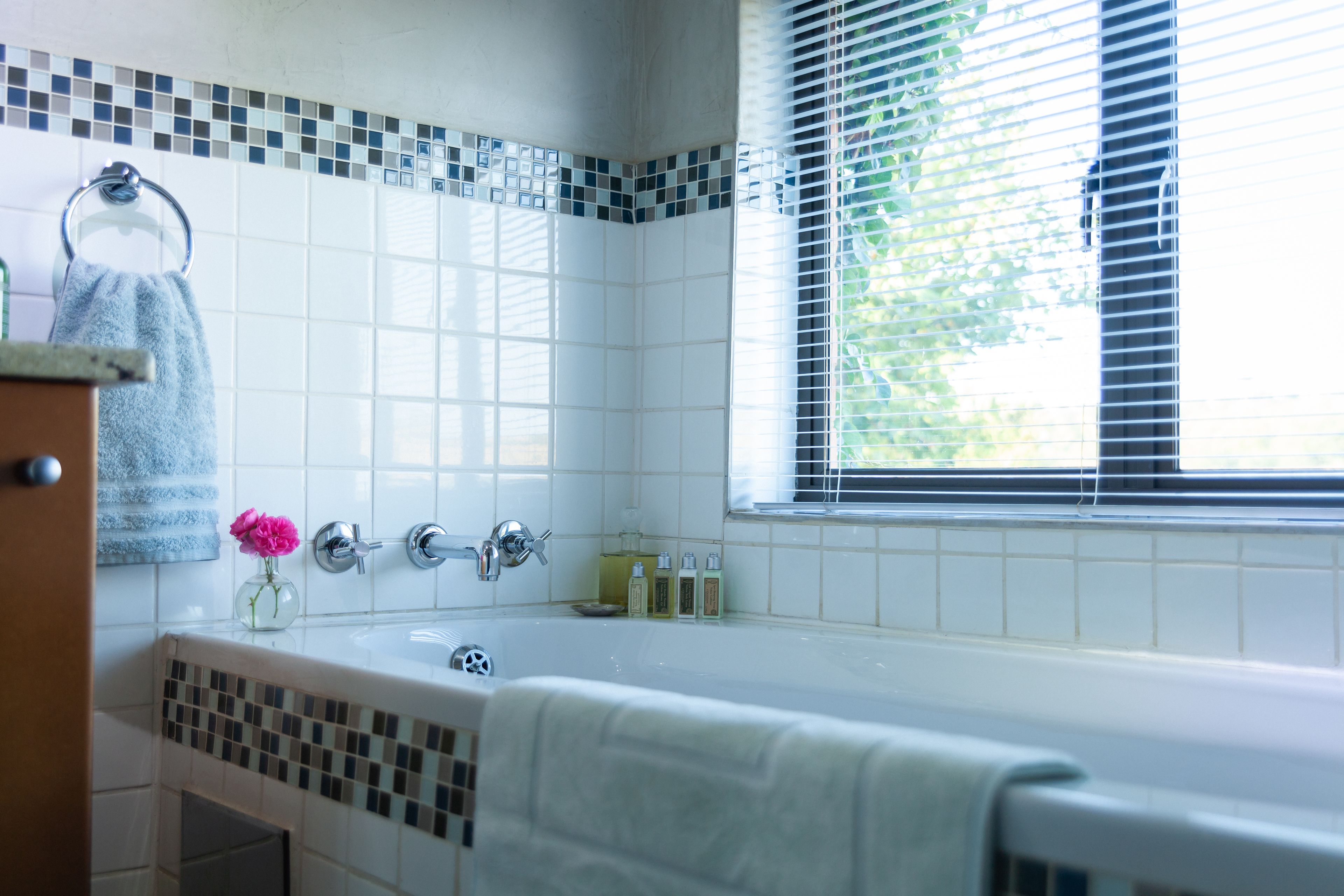 Un baño con bañera, toallero, ventana con persianas y salpicadero de azulejos.