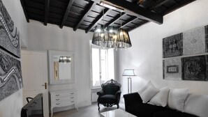 A modern living room with a white sofa, a black armchair, a glass coffee table, and a chandelier. There is a large window with a view of the outside, and artwork on the walls. - Ripa 1 Apartment (Rome)