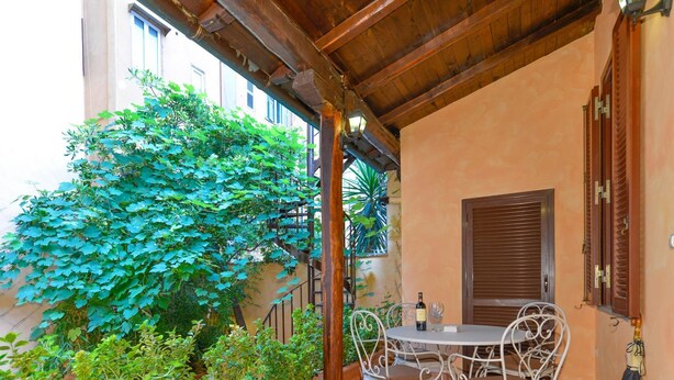 Un porche cubierto con una mesa y sillas, rodeado de plantas en macetas y una pérgola de madera.