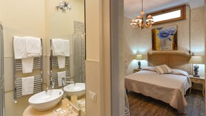 Habitación de hotel con baño que incluye dos lavamanos, un espejo y barras para toallas. El dormitorio tiene una cama, mesitas de noche y un cuadro en la pared.