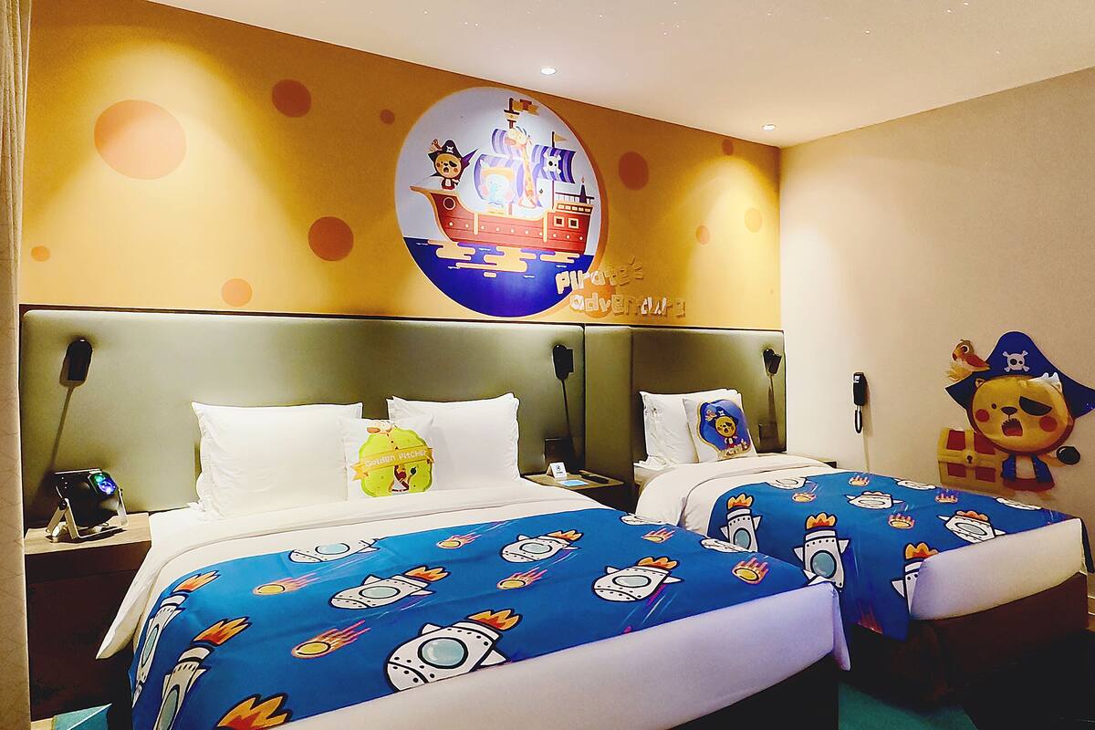 A hotel room with two beds, each with a blue bedspread featuring rocket ship designs. There is a large wall mural of a pirate ship and a golden pitchfork.