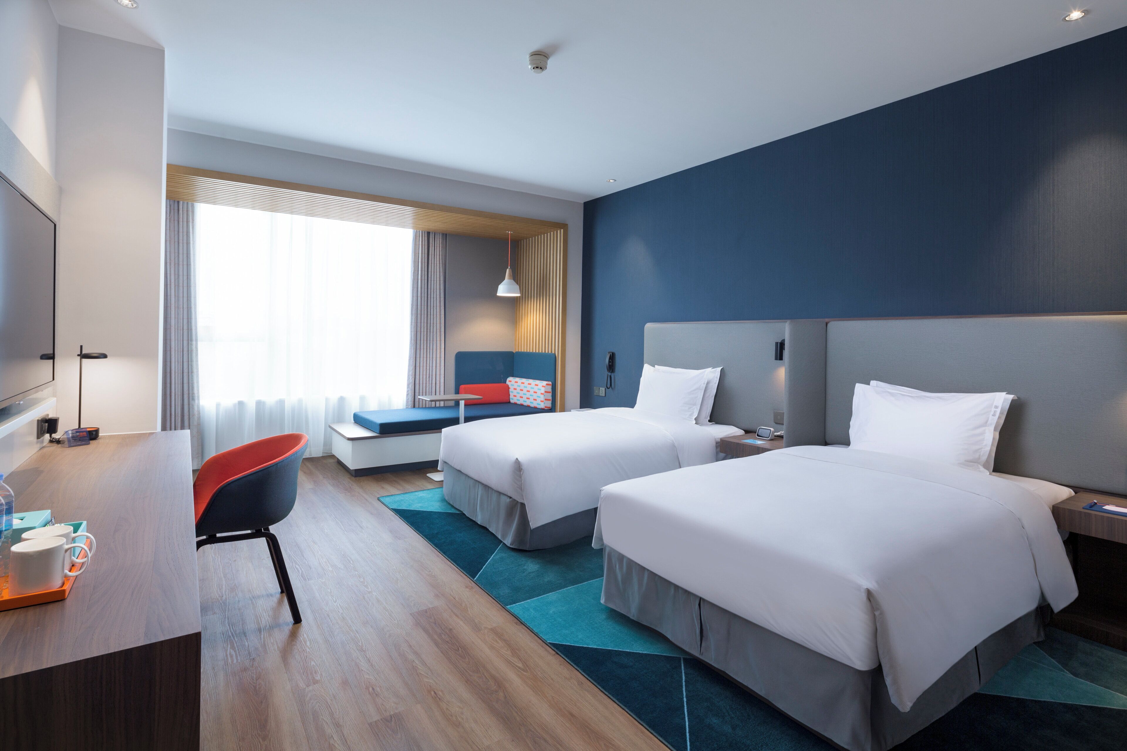 A hotel room with two beds, a desk with a chair, a TV, and a large window with curtains.