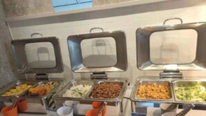 Eine Buffetlinie mit verschiedenen Speisen in Metallbehältern, begleitet von orangefarbenen und weißen Einwegbechern.