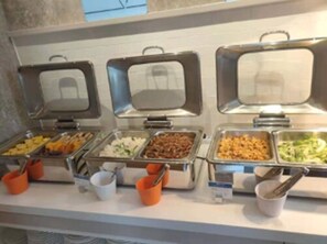Een buffet met verschillende soorten eten in metalen bakken, met oranje en witte wegwerpbekers.