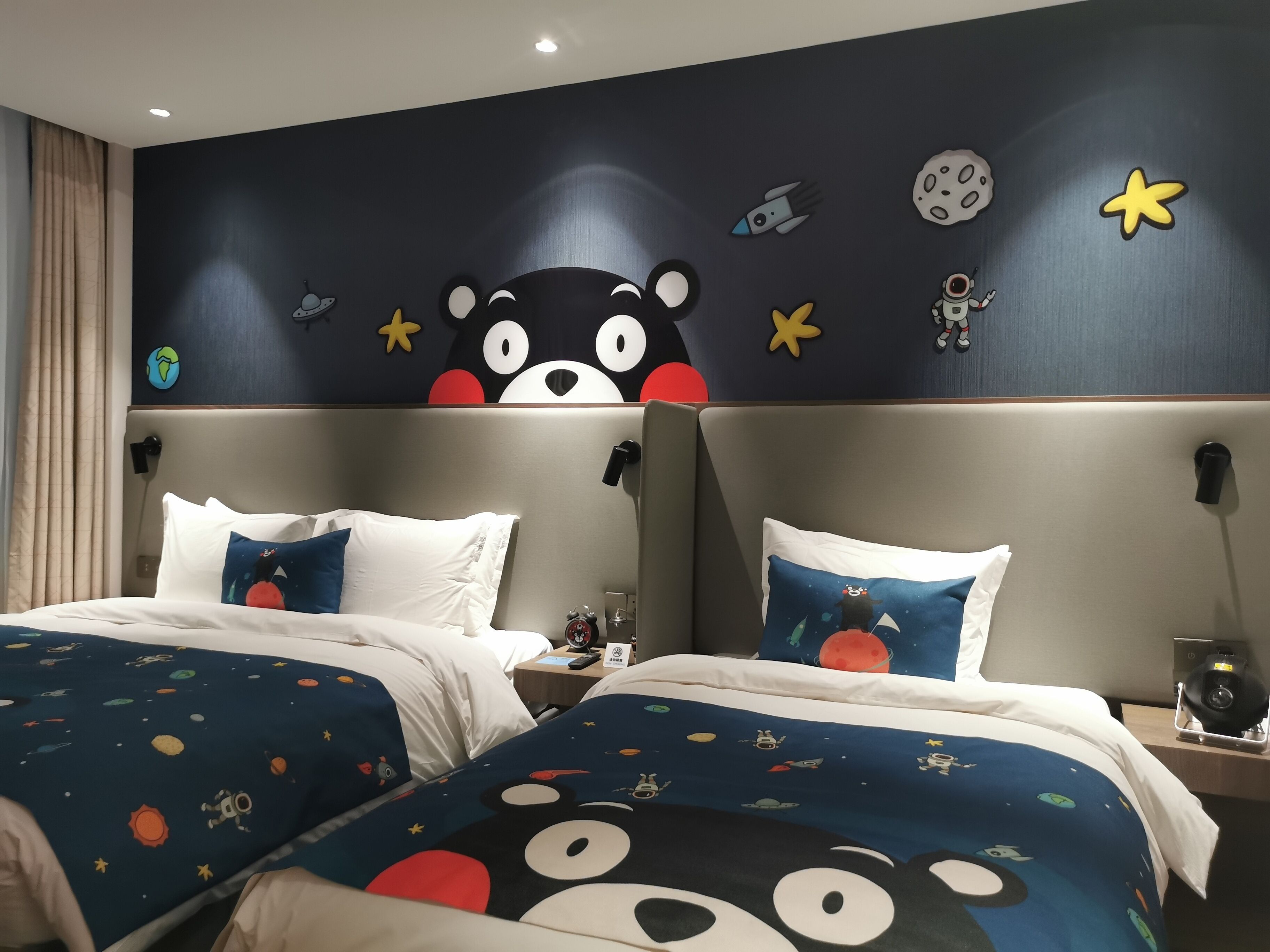 A hotel room with two beds, each with a space-themed bedspread. The room has a dark wall with space-themed decorations, including a bear face and planets. There are two nightstands with lamps and a clock.