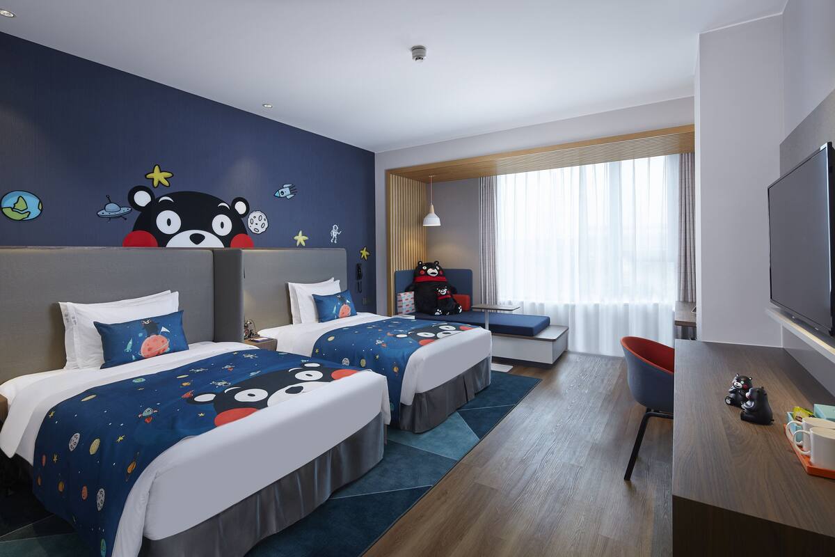 A hotel room with two beds, a desk, a TV, and a large window with curtains.