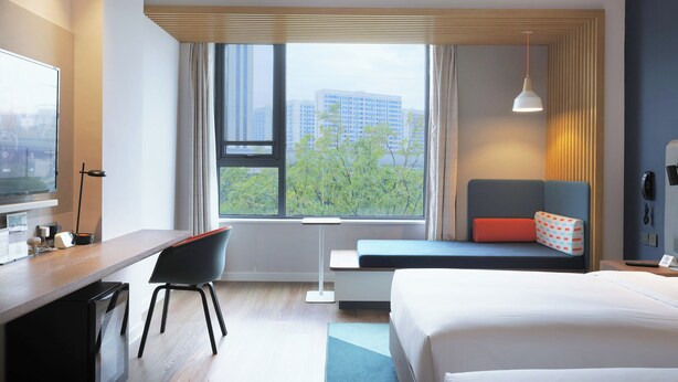 Quarto de hotel moderno com cama, escrivaninha, cadeira, televisão e janela com vista para a cidade.