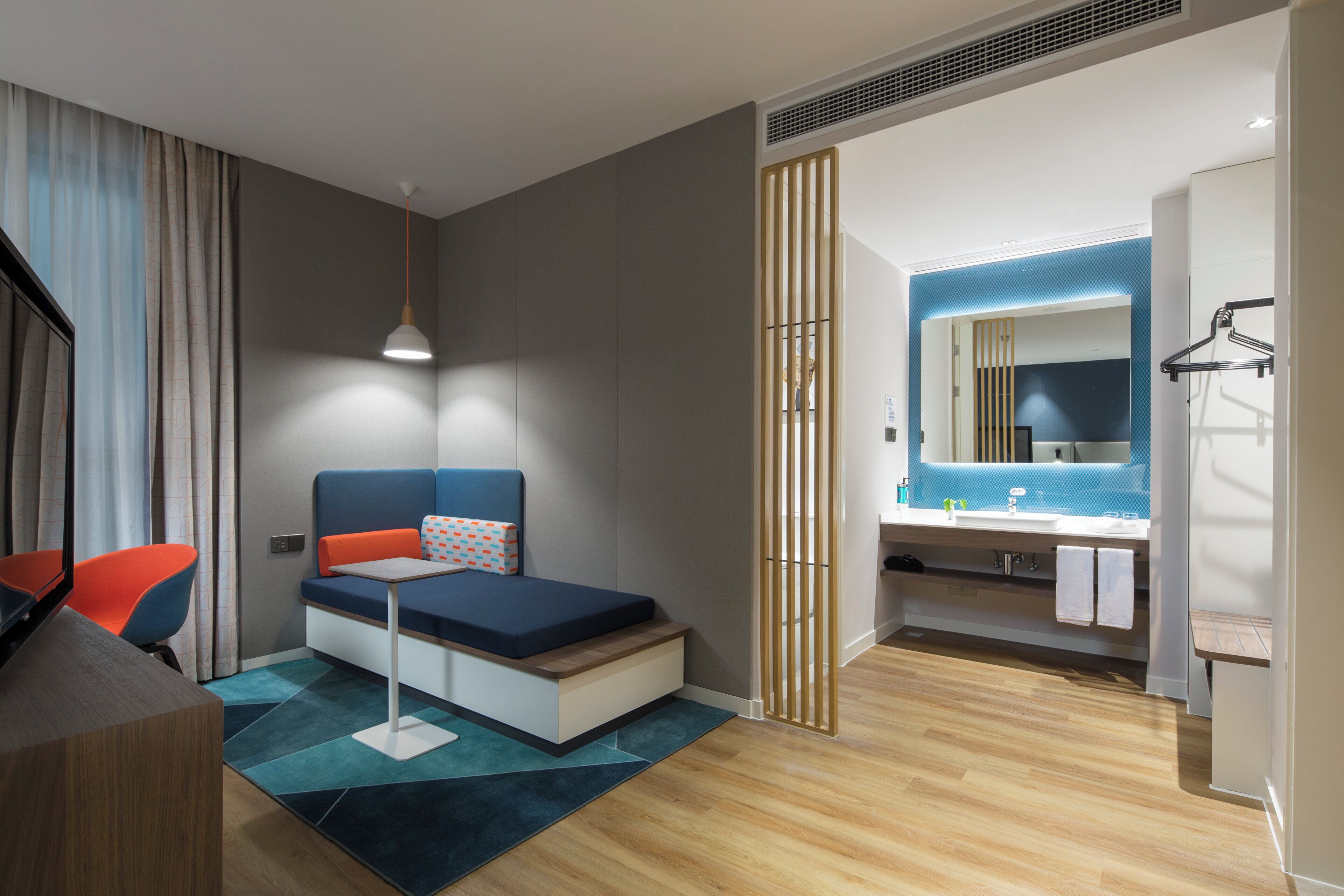 A modern hotel room with a bed, a desk, a chair, a bathroom with a mirror and sink, and a view of the outside.