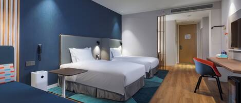 Quarto de hotel com duas camas, uma escrivaninha, uma cadeira, uma mesa pequena e a porta do banheiro.