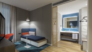 Quarto de hotel moderno com cama, secretária, cadeira e banheiro com espelho e pia.