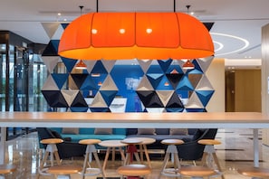 Un interior moderno con un diseño geométrico en la pared, una lámpara colgante naranja en el centro y una zona de estar con mesas y sillas redondas.