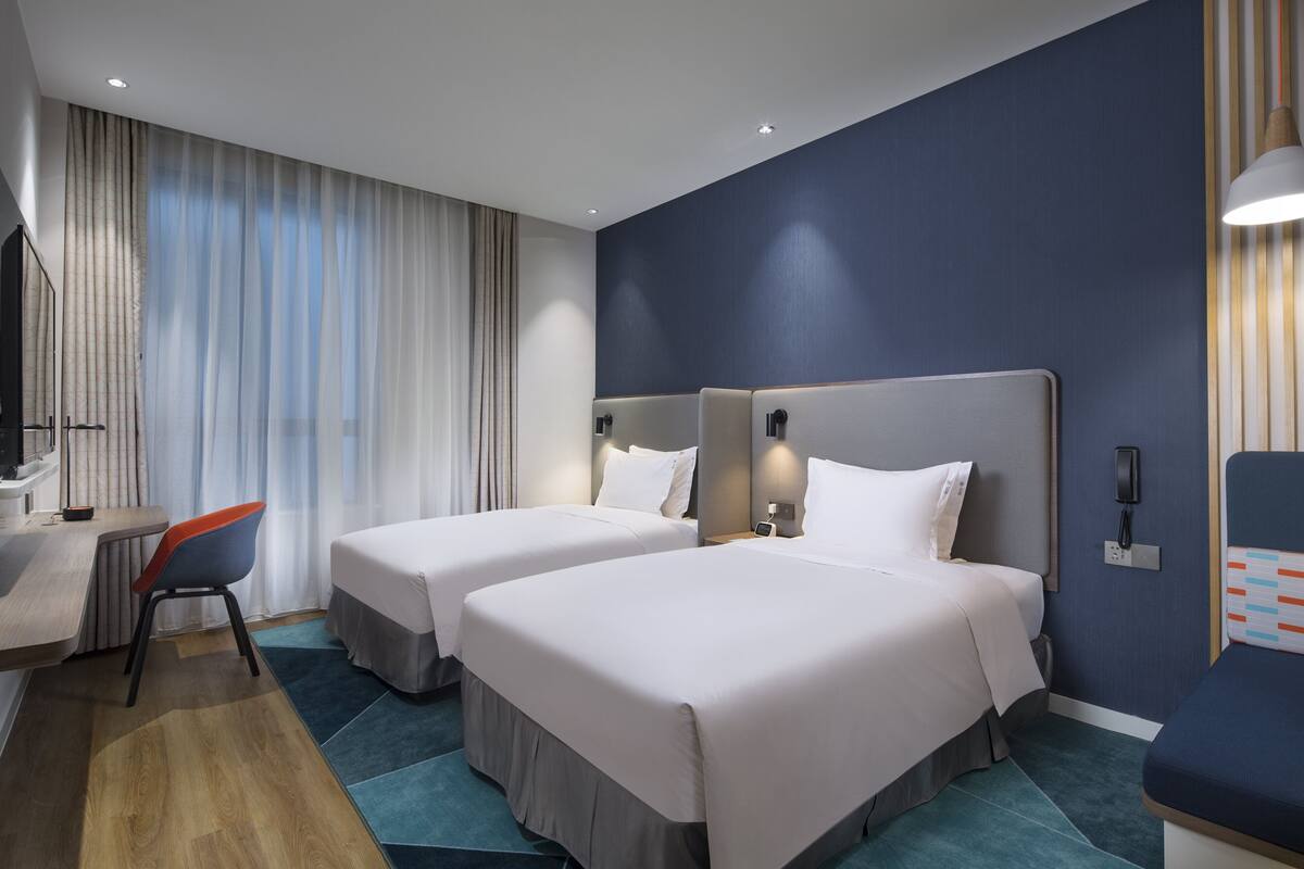A hotel room with two beds, a desk, a chair, and a TV.