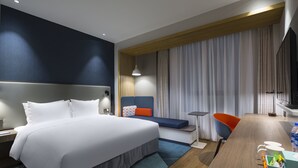Quarto de hotel moderno com uma cama grande, uma escrivaninha com cadeira, uma televisão e uma janela com cortinas.