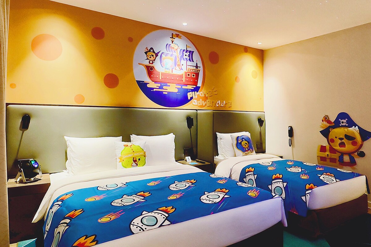 A hotel room with two beds, each with a blue bedspread featuring rocket ship designs. There is a large wall mural of a pirate ship and a golden pitchfork.