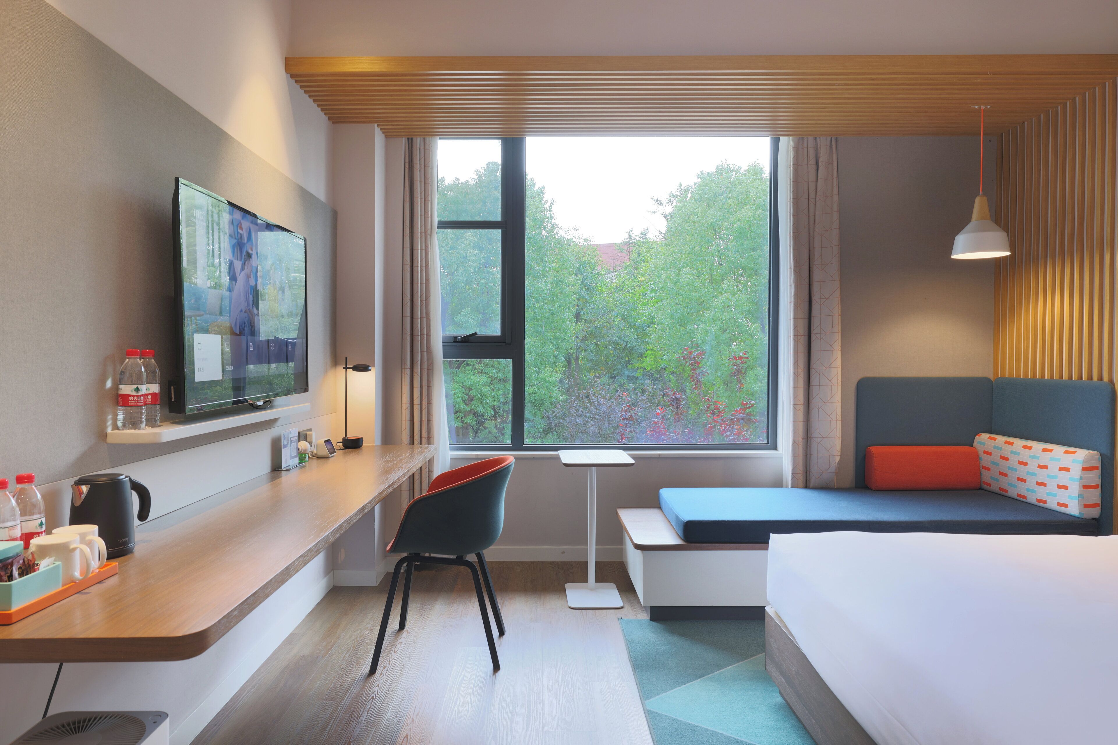 Ein modernes Hotelzimmer mit großem Fenster, einem Flachbildfernseher, einem Schreibtisch mit Stuhl, einem Bett mit blau-orangem Kissen und einem Nachttisch mit Lampe.