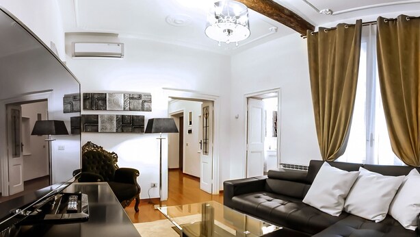 Una sala de estar moderna con un sofá negro, una mesa de centro de vidrio y un televisor. Hay una araña, una lámpara y una puerta que da a otra habitación.