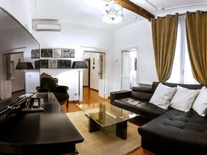 A modern living room with a black sofa, a glass coffee table, and a TV. There is a chandelier, a lamp, and a door leading to another room. - Banchi Apartment (Rome)
