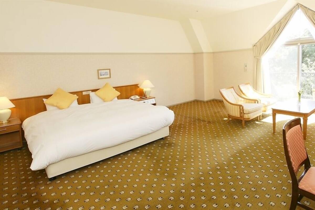 A hotel room with a large bed, two bedside tables with lamps, a small table with chairs, and a large window with curtains.