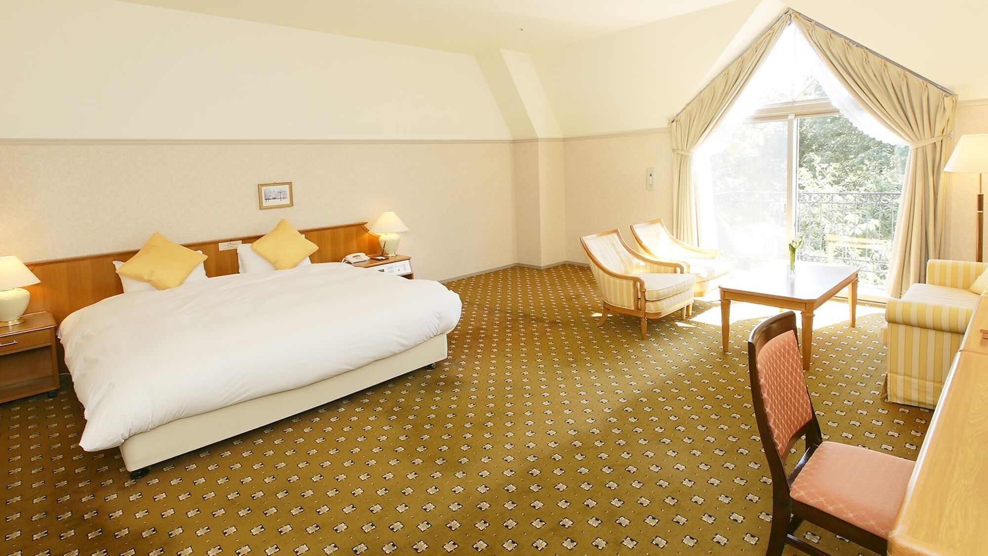 A hotel room with a large bed, two bedside tables with lamps, a small table with chairs, and a large window with curtains.