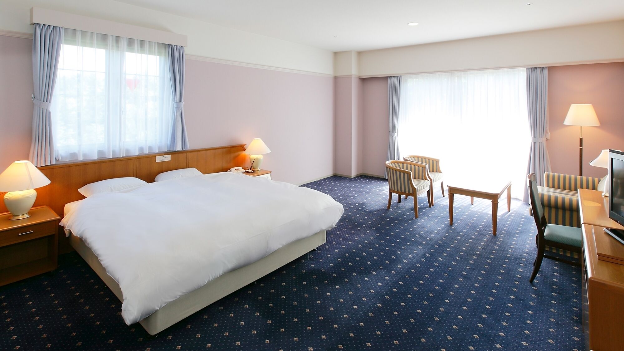 A hotel room with a large bed, two bedside tables with lamps, a desk with a chair, and a television.