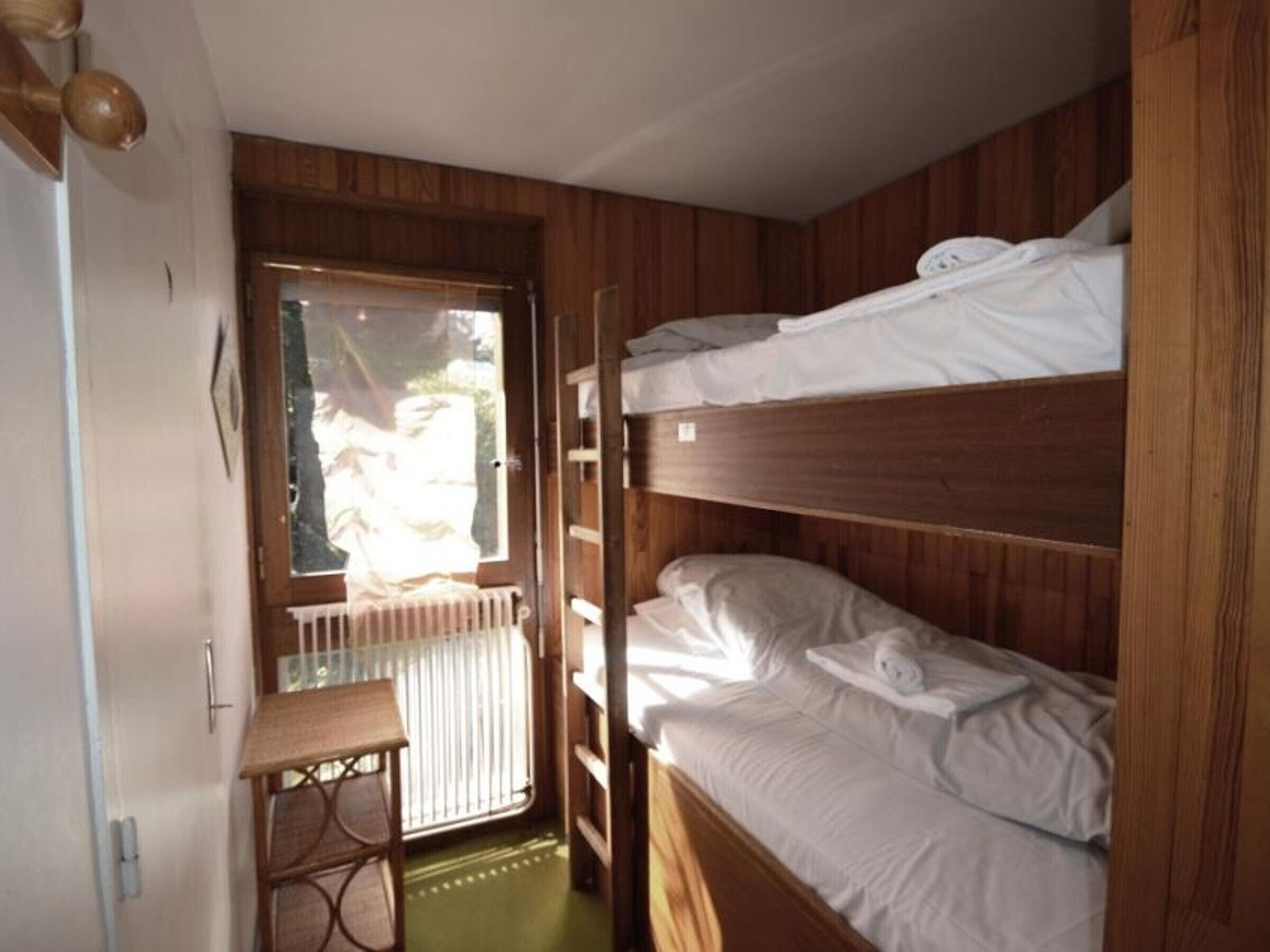 A bunk bed room with wooden walls, a small table, and a window with blinds.
