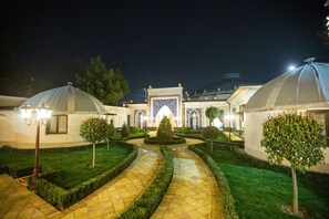 Um pátio com um caminho central, ladeado por gramados e árvores bem cuidados, que leva a um edifício com entrada peculiar e estruturas em forma de cúpula.