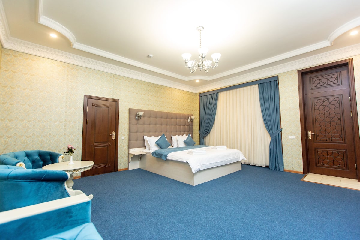 Une chambre d’hôtel avec un grand lit, un canapé bleu, une table ronde avec un vase et deux portes.