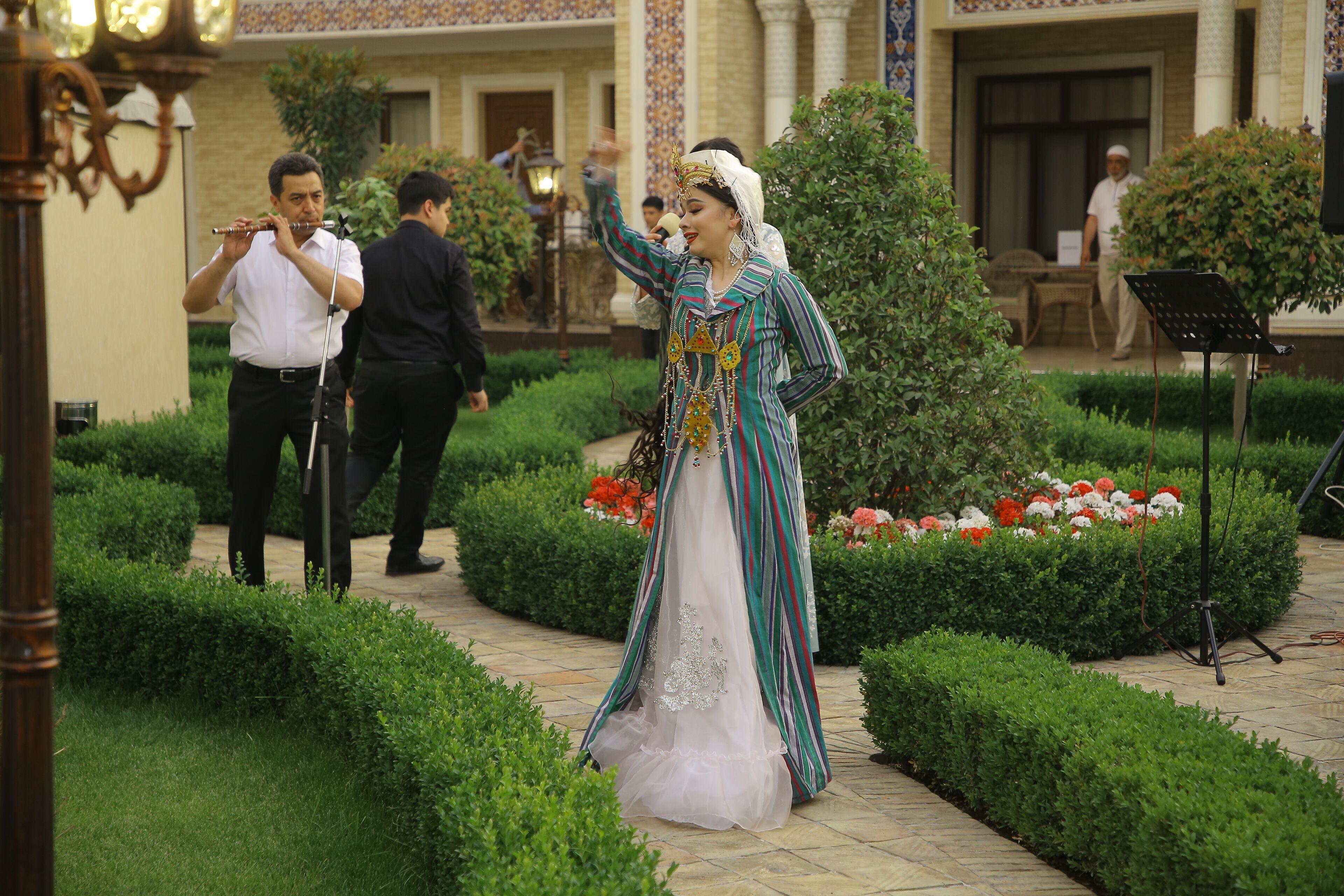 Una mujer vestida con traje tradicional está de pie en un jardín, cerca hay un soporte para micrófono y un atril musical.