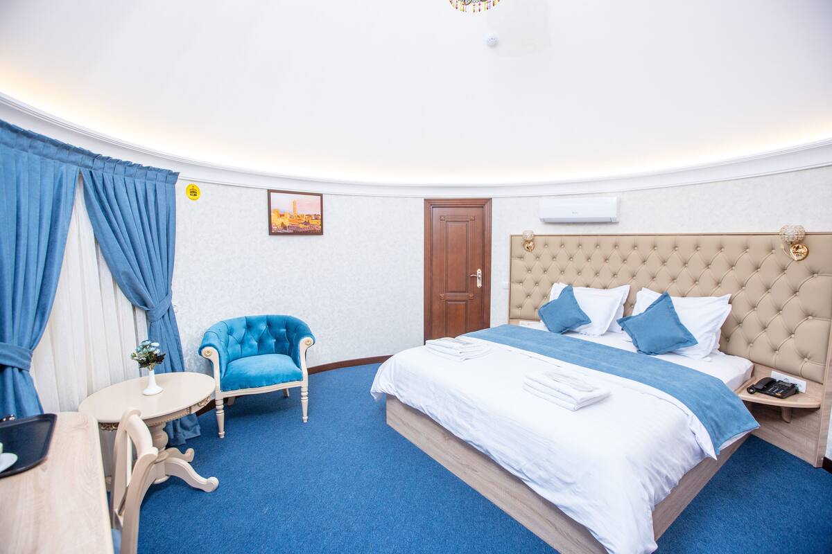 Une chambre d’hôtel avec un grand lit, un fauteuil bleu, une petite table ronde et une table de chevet avec un téléphone.